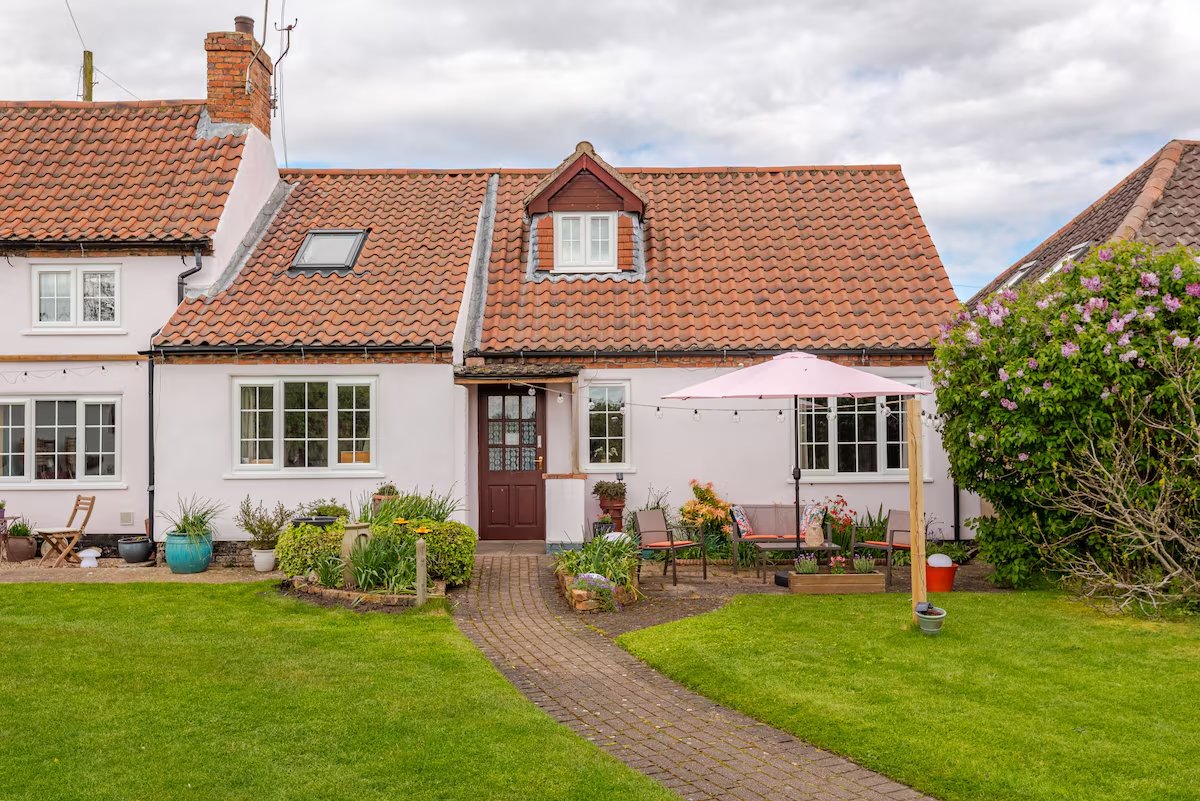 A quaint house with a red-tiled roof, pastel pink walls, and a wooden front door. The yard includes a seating area with a pink umbrella and a well-manicured lawn with flower beds.
