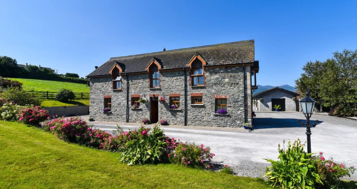 A picturesque stone house set against a backdrop of lush greenery and a clear blue sky. The house has two stories and is made of gray stone with a dark gray shingled roof.  
A well-maintained garden with vibrant pink and red flowers borders the gravel driveway leading to the house. 