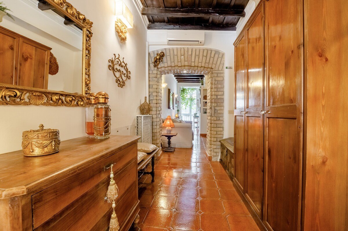 A well-lit, narrow hallway with a warm, rustic decor.  terracotta tile floor, wooden dresser and wardrobe, gold-framed mirror, ornate decorations, and a view of a living area with a sofa and balcony.

The hallway extends to a living area visible in the background, furnished with a white sofa, a small wooden table with an orange lampshade, and a balcony door framed with green plants.