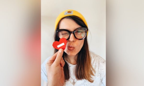 Female wearing yellow beanie and black glasses, making kissing gesture towards a red lego heart someone is holding up
